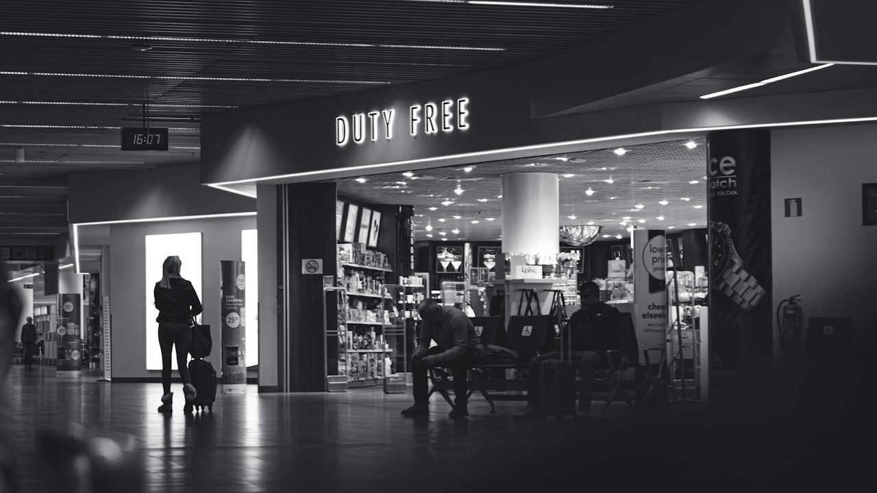 A duty-free store front in an airport terminal, lettering above the entrance and a passenger walking past with a suitcase